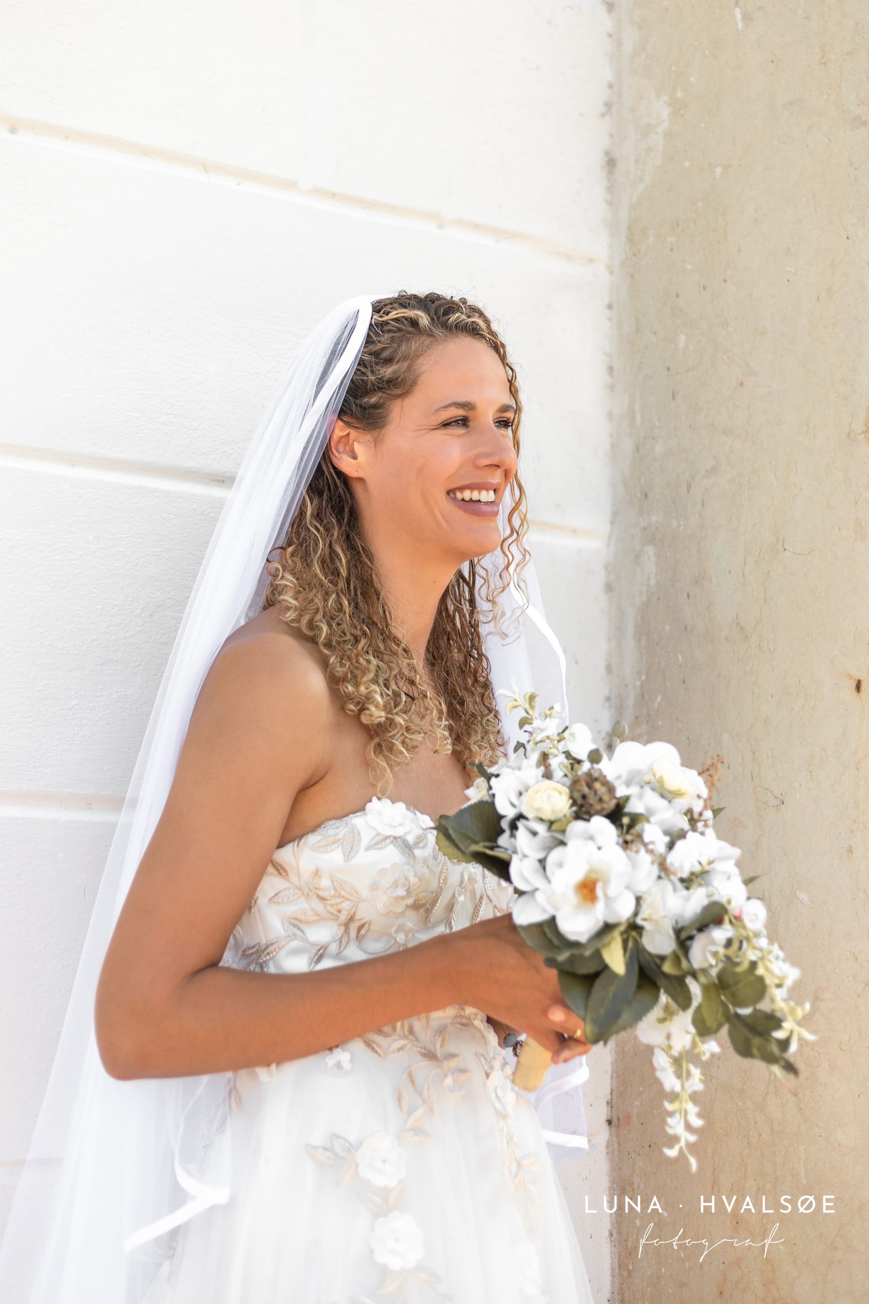Brud smiler med slør og hvide brudebuket foran Hørsholm Kirke