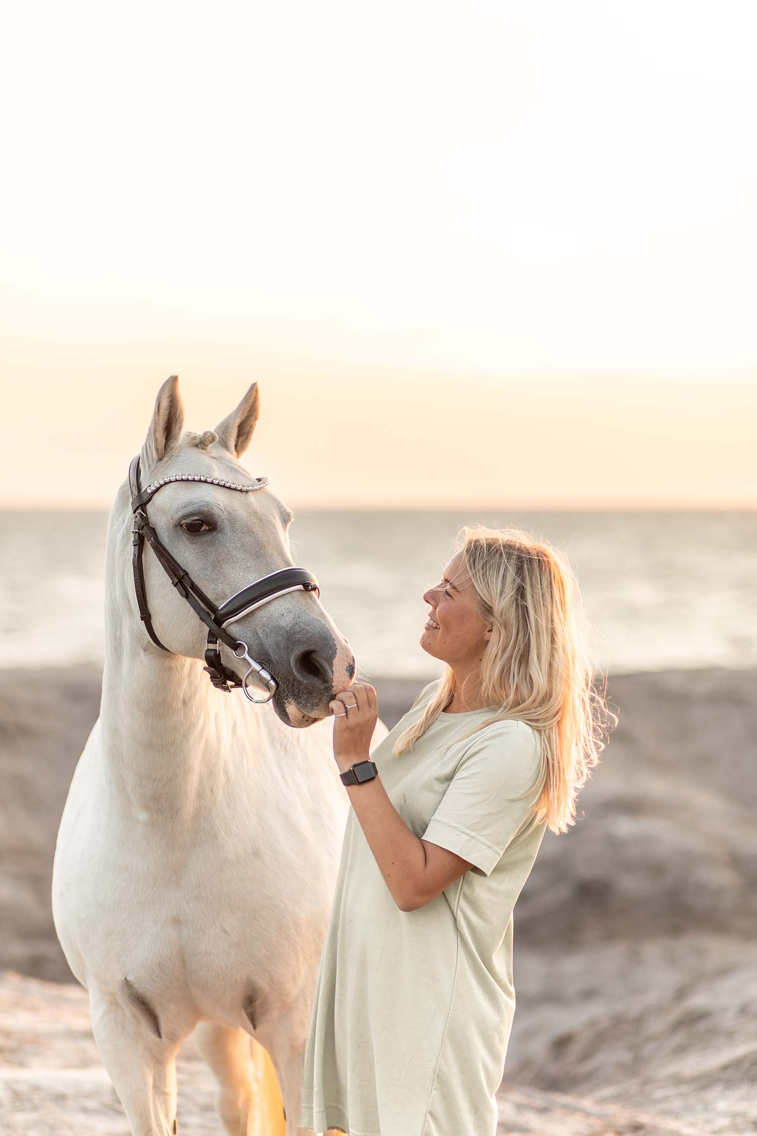 hvid pony og pige står i solnedgangen ved strand